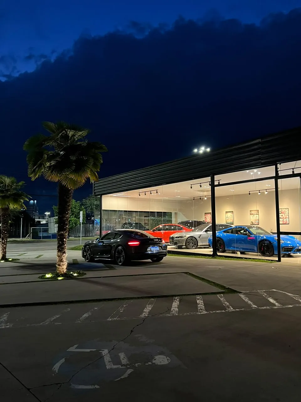 Cenci Motors ao anoitecer, com a vitrine acesa, palmeiras e um Porsche preto na entrada