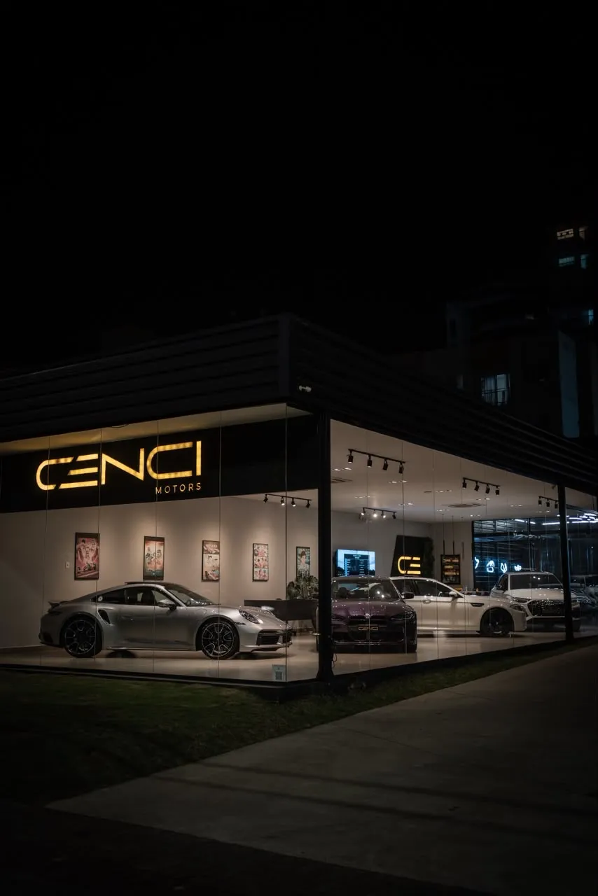 Fachada da Cenci Motors à noite, com o letreiro dourado aceso e um Porsche prata na vitrine